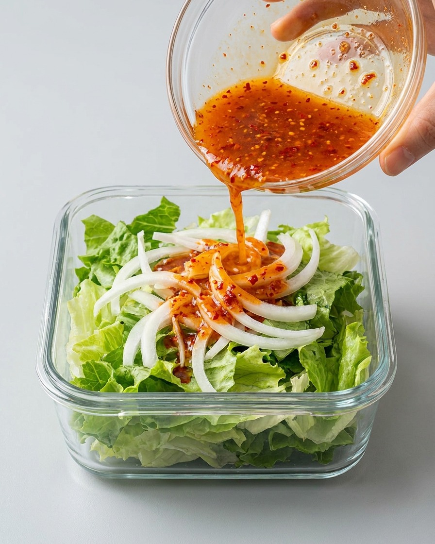 Vertiendo un caldo de kimchi rojo y picante desde un bol pequeño sobre lechuga y cebolla en un recipiente rectangular.