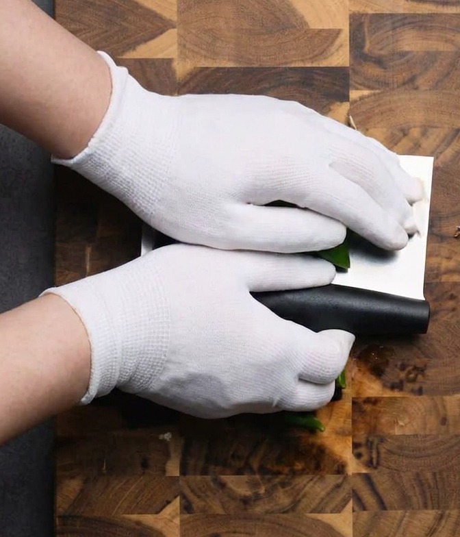 Manos con guantes usando un hacha de cocina para picar chiles verdes sobre una tabla de madera.