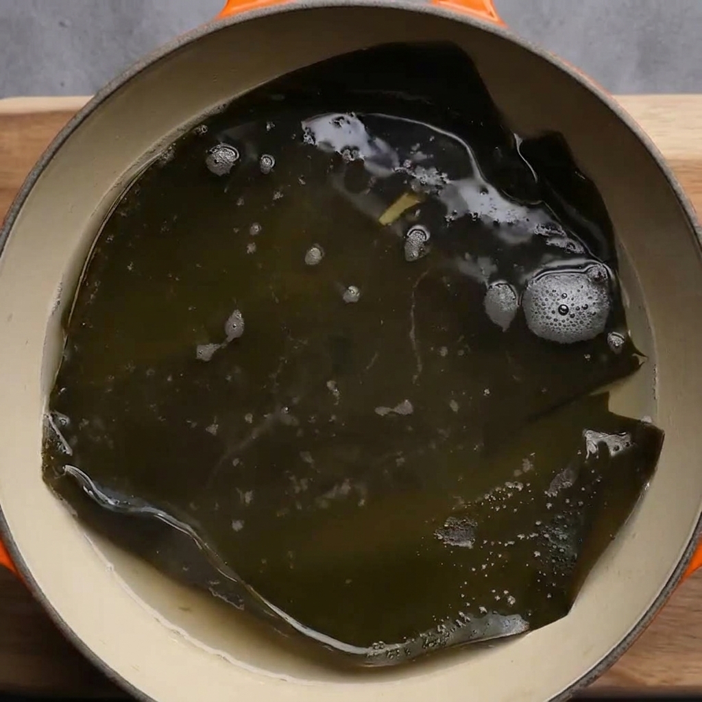 Un trozo grande de alga kombu verde oscuro hirviendo a fuego lento en una olla con agua burbujeante.