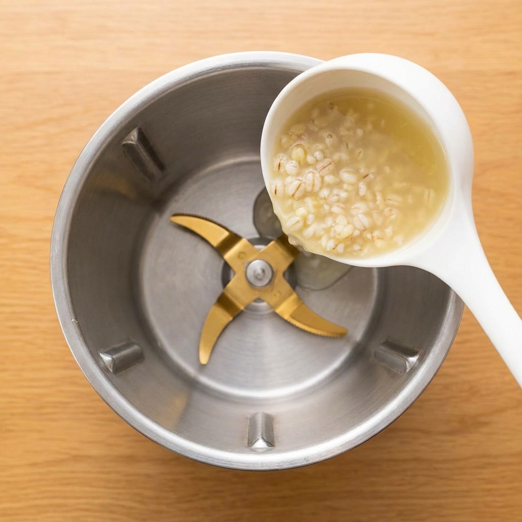 Un cucharón blanco vertiendo gachas de cebada espesas y pálidas en el vaso de una licuadora de acero inoxidable.