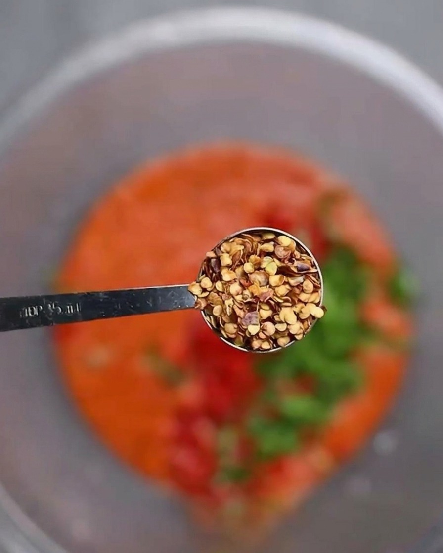 Una cuchara medidora con semillas de chile secas sostenida sobre un bol con pasta de chile roja y vegetales picados.