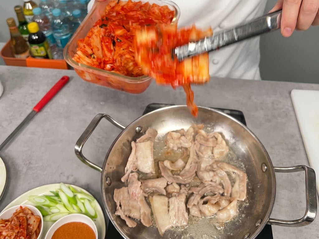 Una porción generosa de kimchi rojo brillante pasando de un recipiente de vidrio a la sartén con panceta parcialmente cocida.