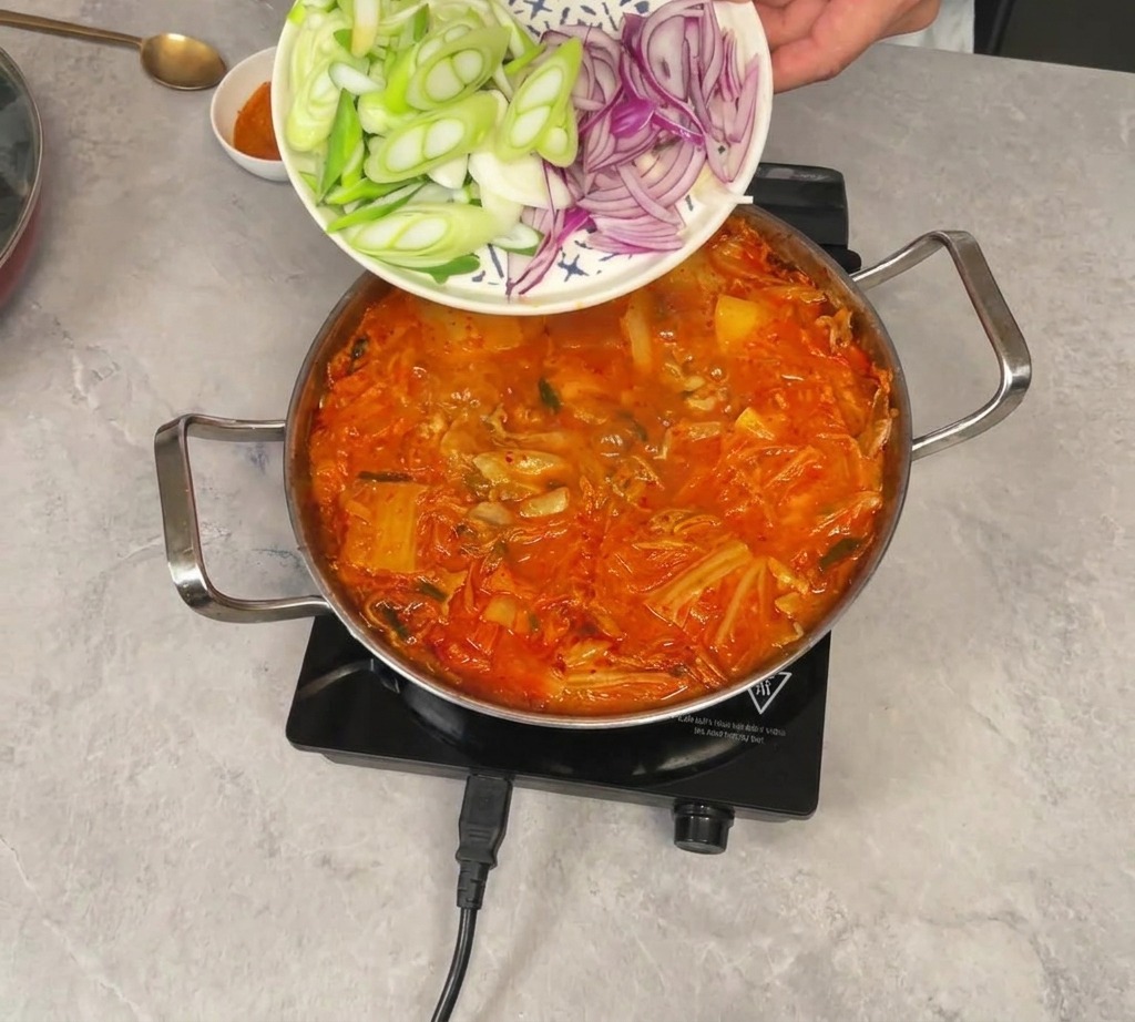 Cebollín y cebolla morada rebanados deslizándose desde un plato hacia la olla con el estofado de kimchi.