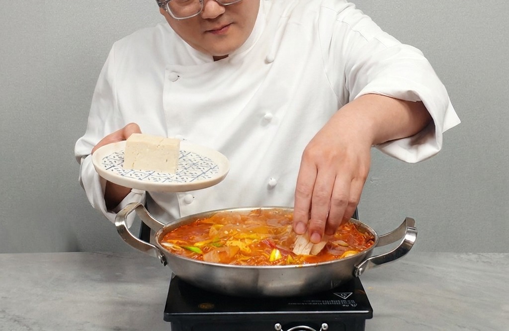 Una persona colocando rodajas de tofu blanco firme en una olla de estofado de kimchi hirviendo.