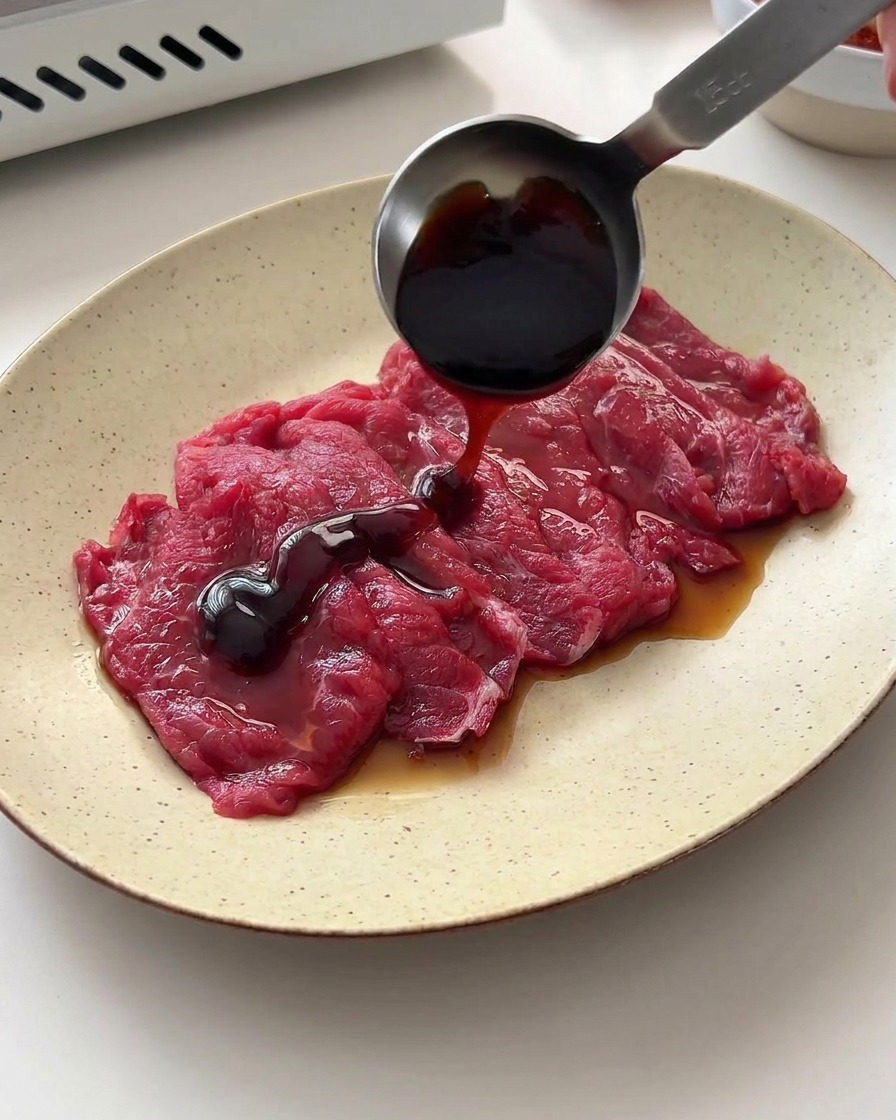 Salsa de soja y salsa de ostras vertiéndose sobre la ternera cruda con una cuchara medidora.