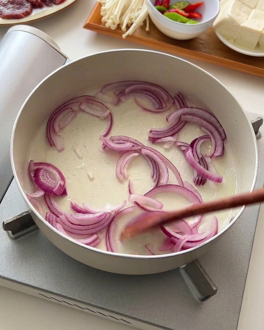 Cebolla morada laminada removiéndose con un utensilio de madera en una sartén blanca poco profunda.