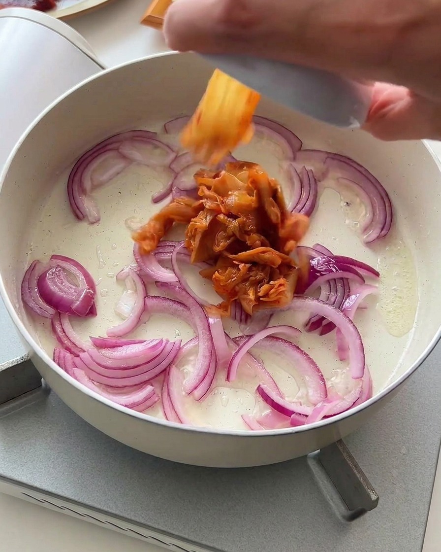 Kimchi troceado cayendo en una sartén con cebolla morada sofriéndose suavemente.