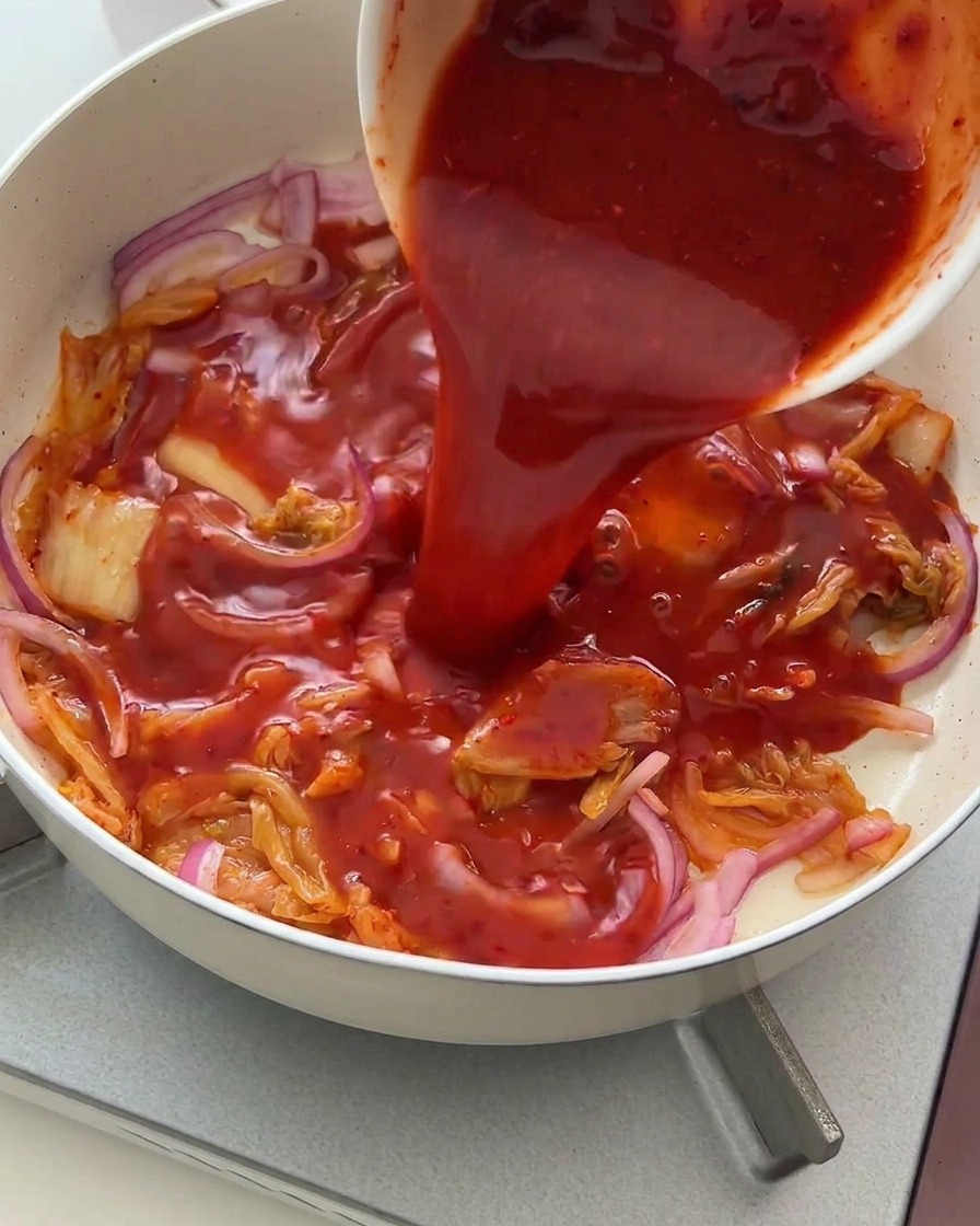 Salsa roja vibrante vertiéndose desde un cuenco sobre el sofrito de kimchi y cebolla.