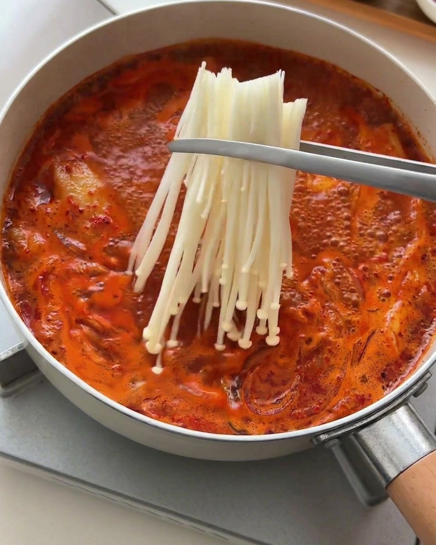 Pinzas metálicas colocando un manojo de setas enoki frescas en un caldo rojo burbujeante.
