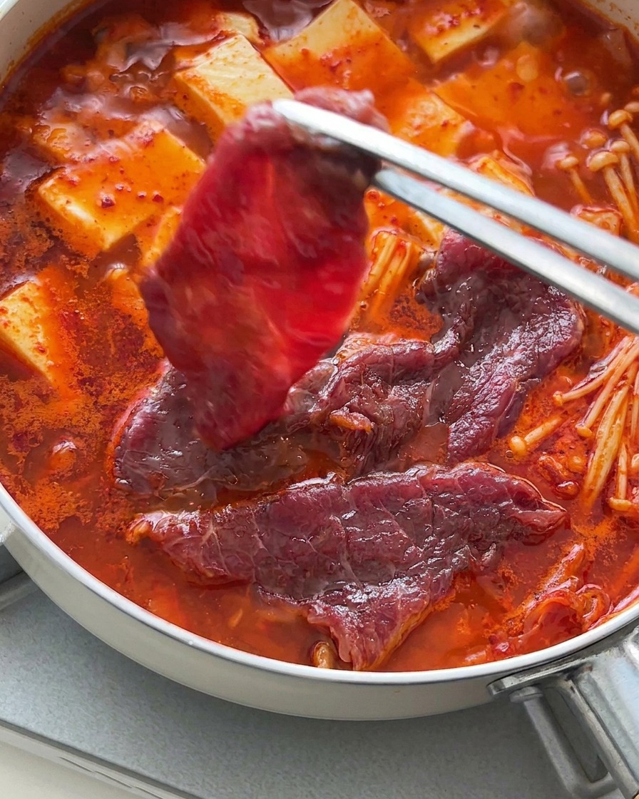 Pinzas colocando láminas finas de ternera cruda sobre el estofado burbujeante de tofu y kimchi.