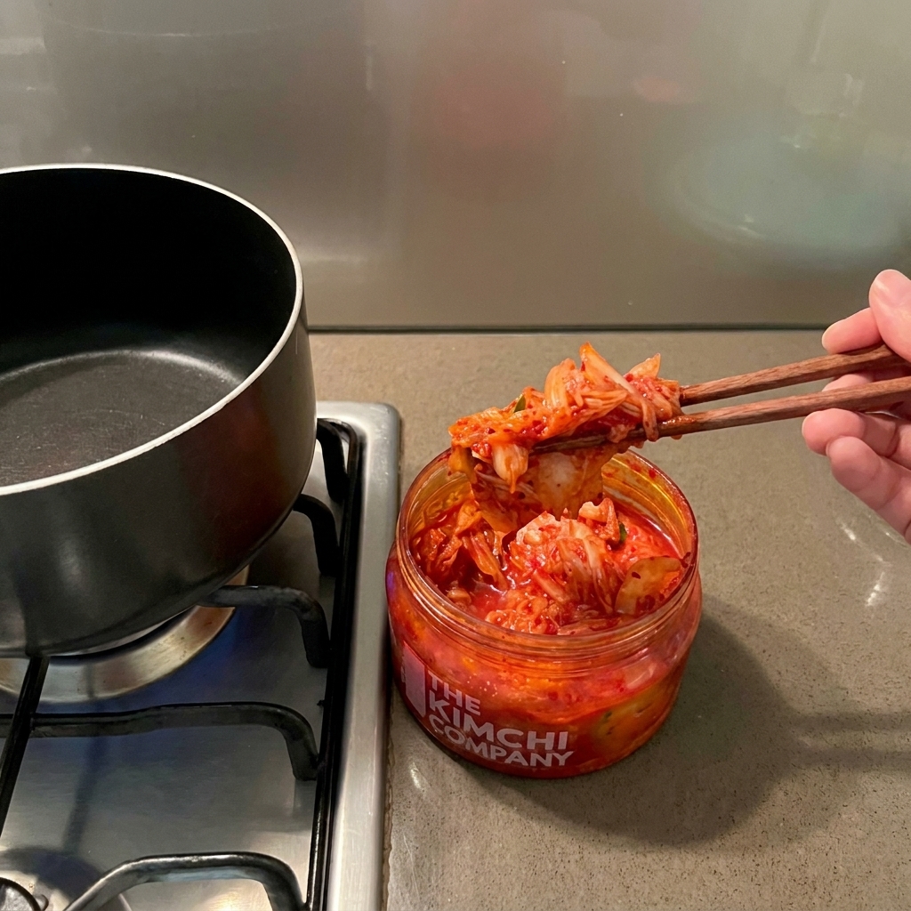 Una mano usando palillos de madera para pasar kimchi rojo de un frasco a una olla de cocina vacía.