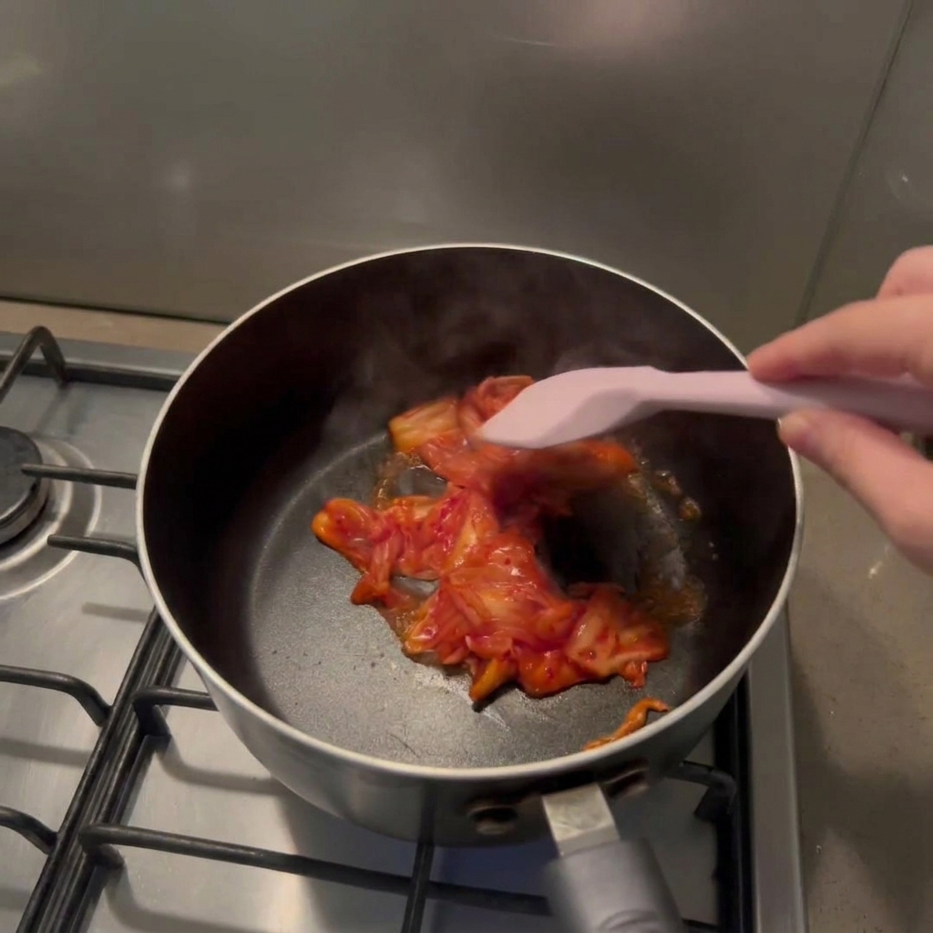 Una mano usando una espátula de silicona para saltear trozos de kimchi rojo dentro de una olla negra.