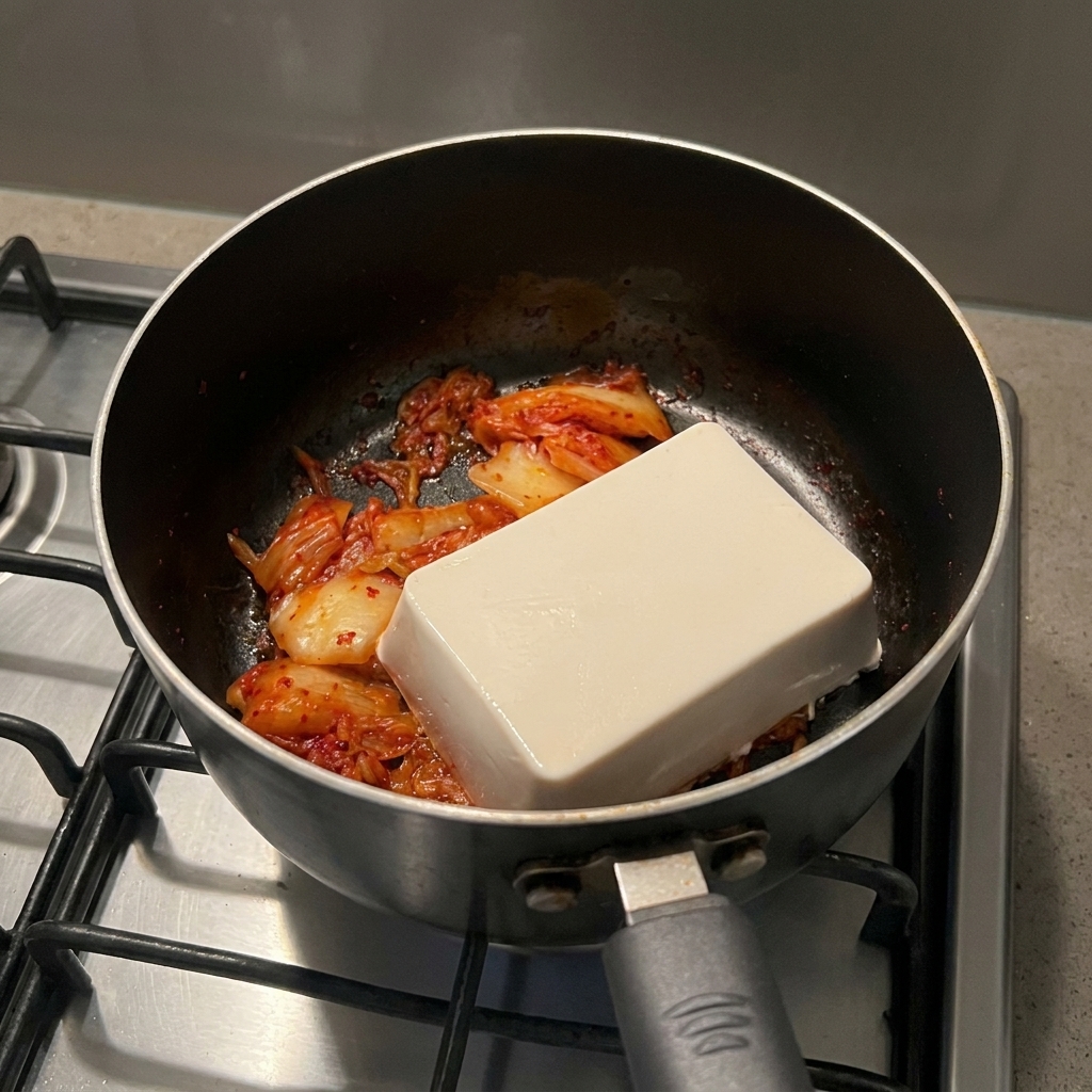 Un bloque rectangular de tofu blanco suave sobre una base de kimchi rojo cocinado en una olla.