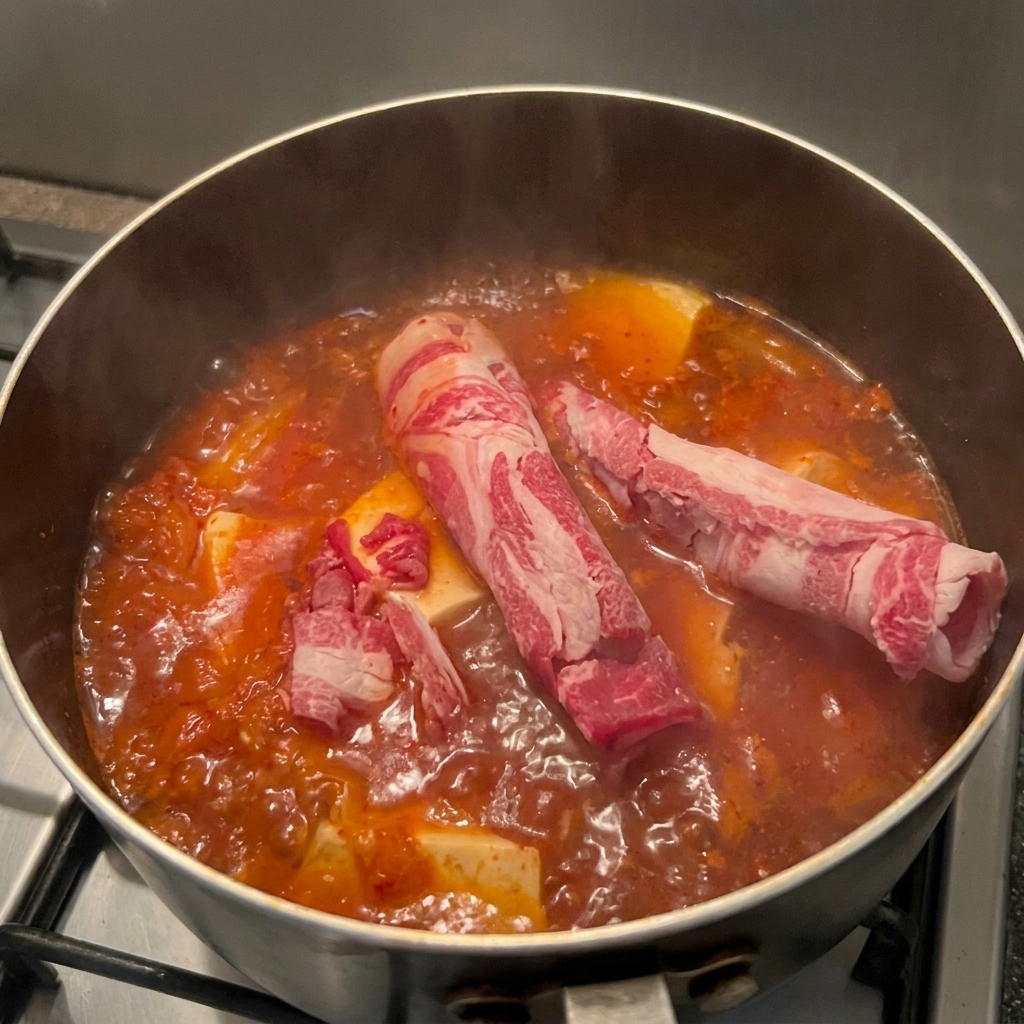 Láminas finas de ternera cruda veteada introduciéndose en una olla de sopa de kimchi hirviendo con tofu.