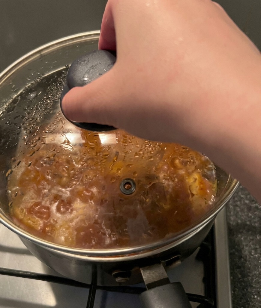 Una mano colocando una tapa de cristal sobre una olla de sopa de kimchi humeante.