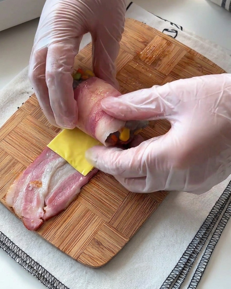 Manos enrollando cuidadosamente una tira de bacon alrededor del relleno de gambas y queso.