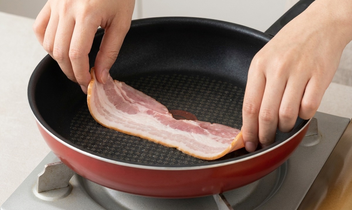 Manos colocando tiras de tocino crudo en una sartén precalentada con un poco de aceite.