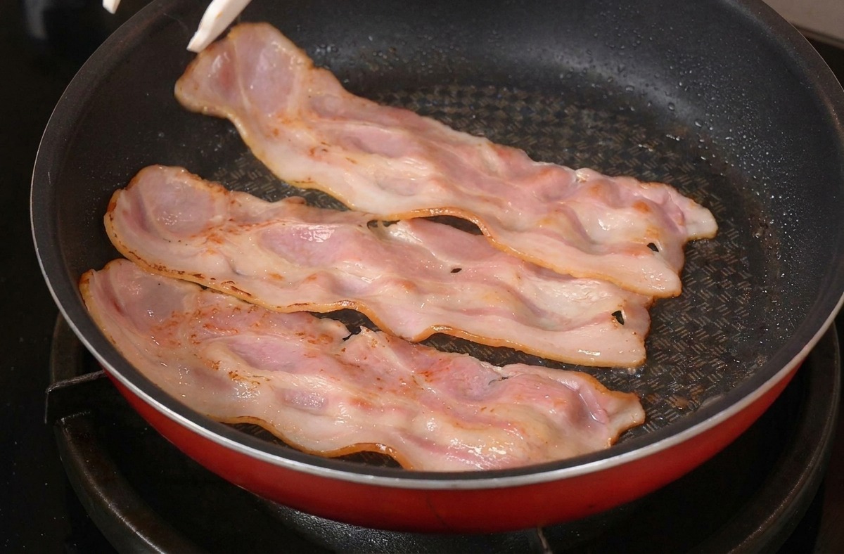 Pinzas volteando tiras de tocino parcialmente cocidas en una sartén roja sobre la estufa.