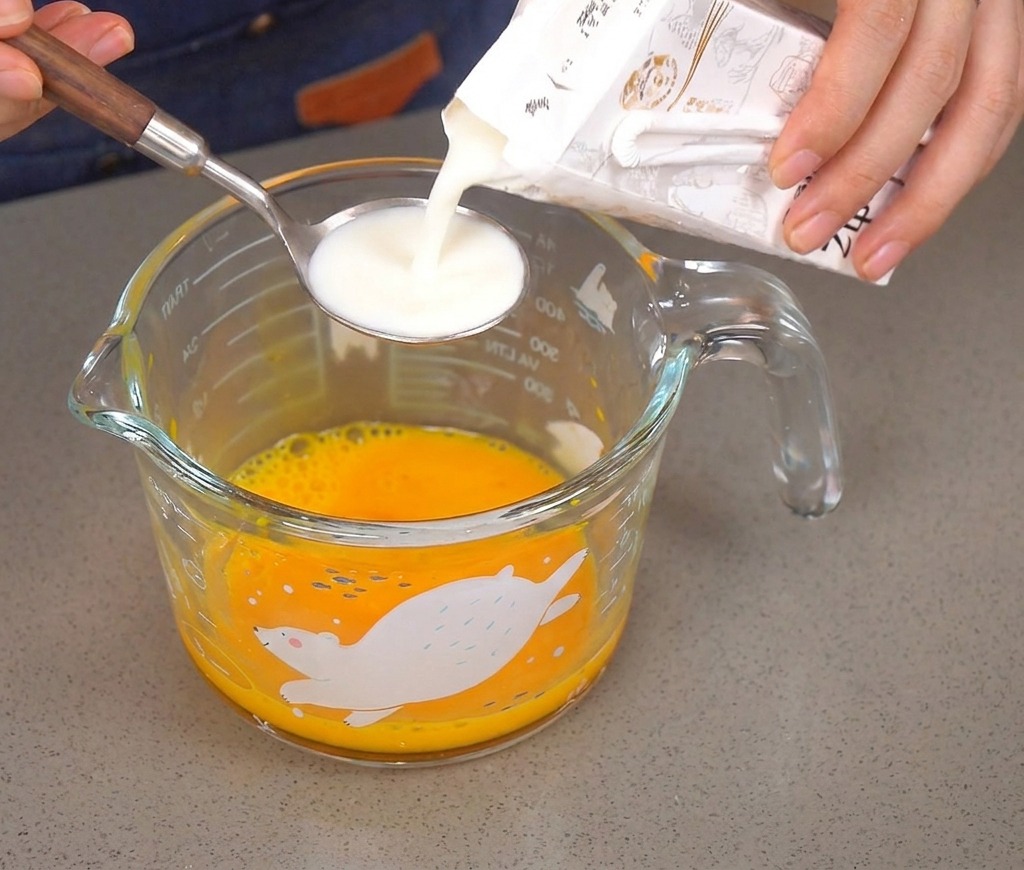 Vertiendo leche desde un cartón en una cuchara de plata sobre un cuenco con huevos batidos.