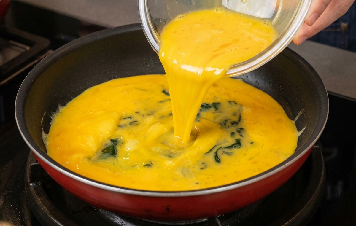 Mezcla de huevos batidos vertiéndose desde un cuenco de vidrio a una sartén limpia.