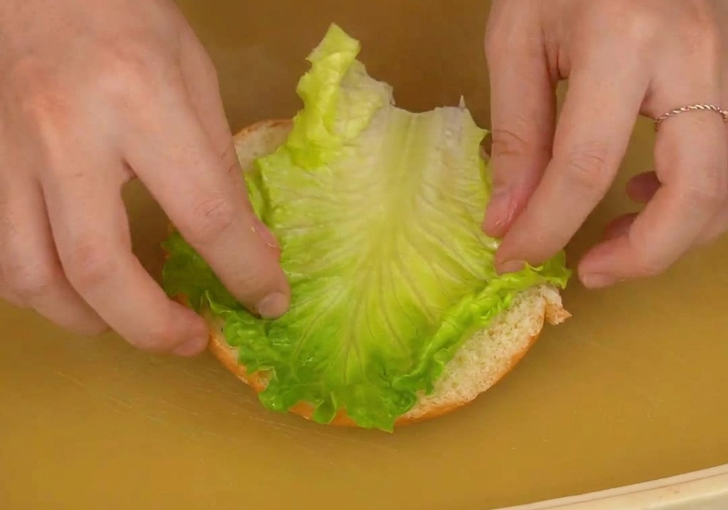 Dos manos colocando una hoja de lechuga fresca sobre la mitad inferior de un pan de hamburguesa.