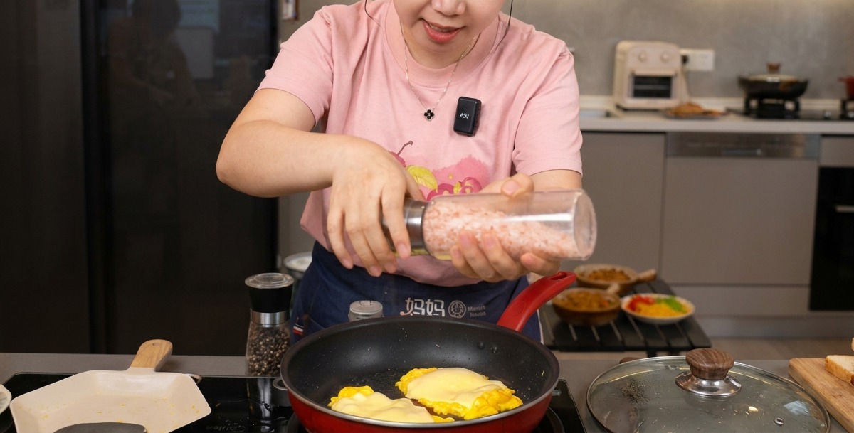 Una persona moliendo sal marina y pimienta negra sobre huevos revueltos con queso fundido en una sartén roja.