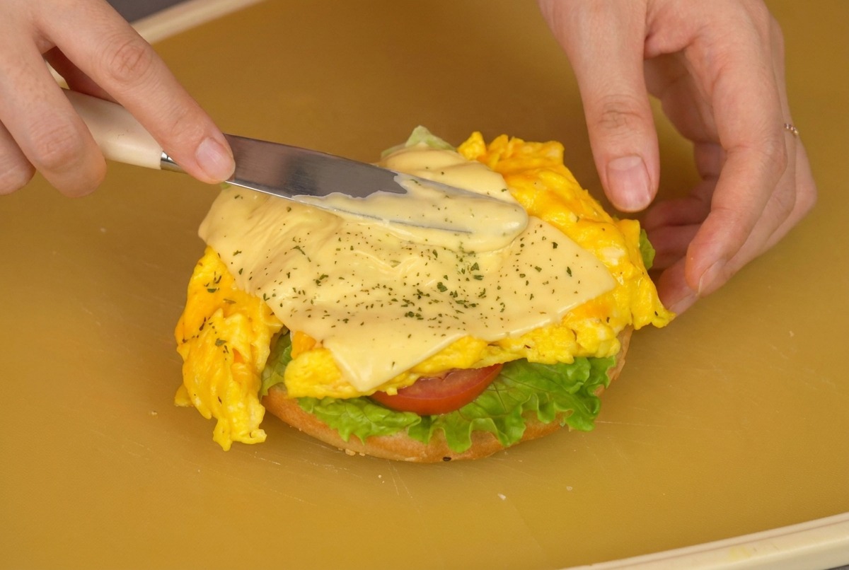 Primer plano de una persona usando una espátula para colocar el huevo con queso sobre el pan con lechuga y tomate.