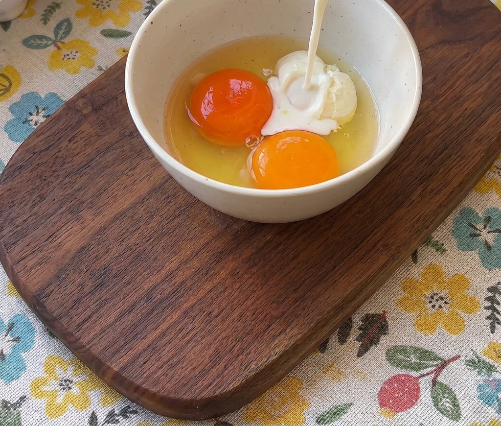 Leche vertiéndose desde un vaso en un bol con dos yemas de huevo de color naranja intenso.
