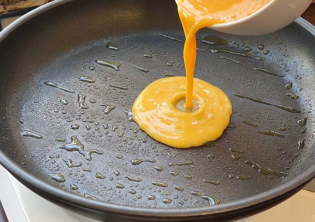 Mezcla de huevo batido vertiéndose desde un bol blanco a una sartén negra con aceite caliente.