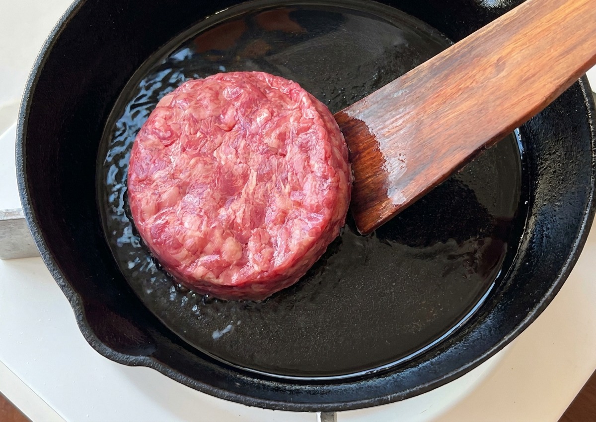 Una hamburguesa de ternera cruda colocándose en una sartén de hierro fundido caliente.