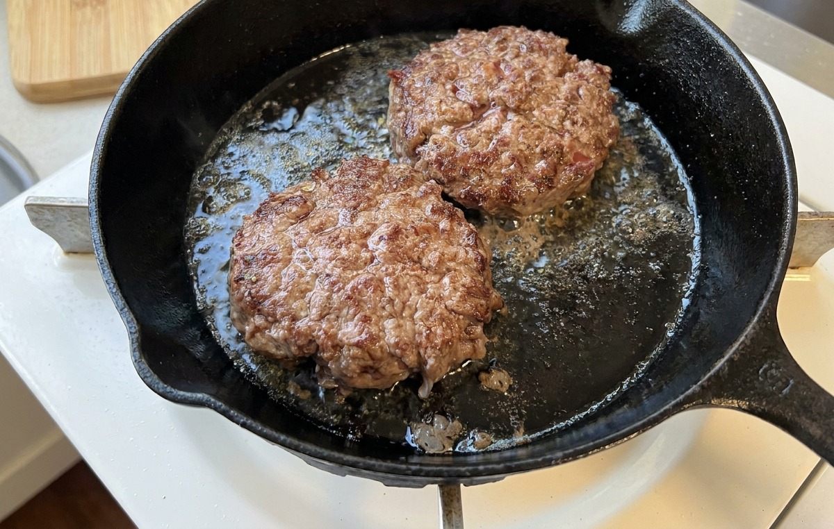 Dos hamburguesas de ternera chisporroteando en una sartén con una costra bien caramelizada.
