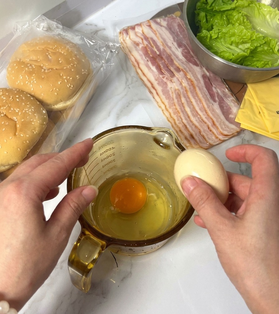 Una persona rompiendo un huevo crudo en una jarra medidora de cristal que ya contiene una yema y clara, con ingredientes de hamburguesa al fondo.