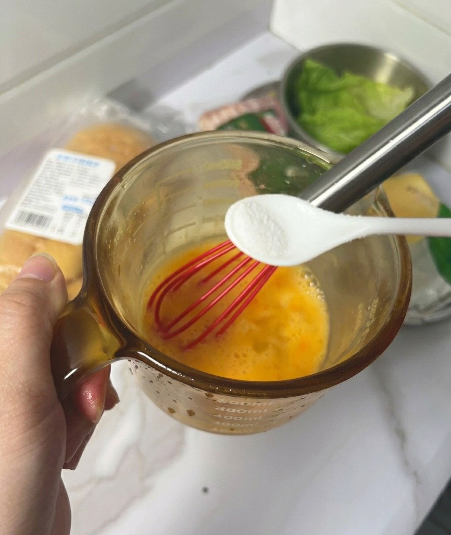 Una mano sosteniendo una pequeña cuchara blanca con sal sobre una taza de cristal con huevos amarillos batidos y un batidor rojo.