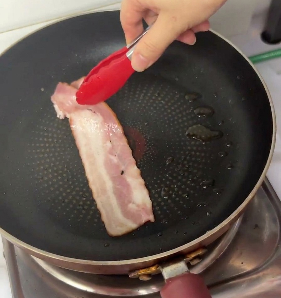 Una mano usando pinzas de punta roja para colocar una tira larga de tocino rosado crudo en una sartén antiadherente negra.