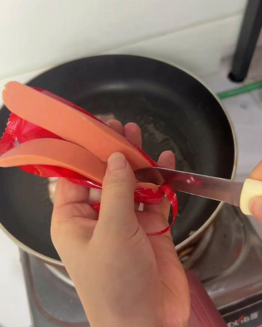 Una persona usando un cuchillo pequeño para cortar una salchicha de hot dog por la mitad a lo largo sobre una sartén.