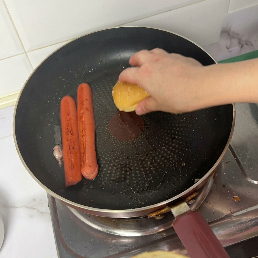 Una persona colocando la parte superior de un pan de sésamo boca abajo en una sartén junto a dos salchichas dorándose.
