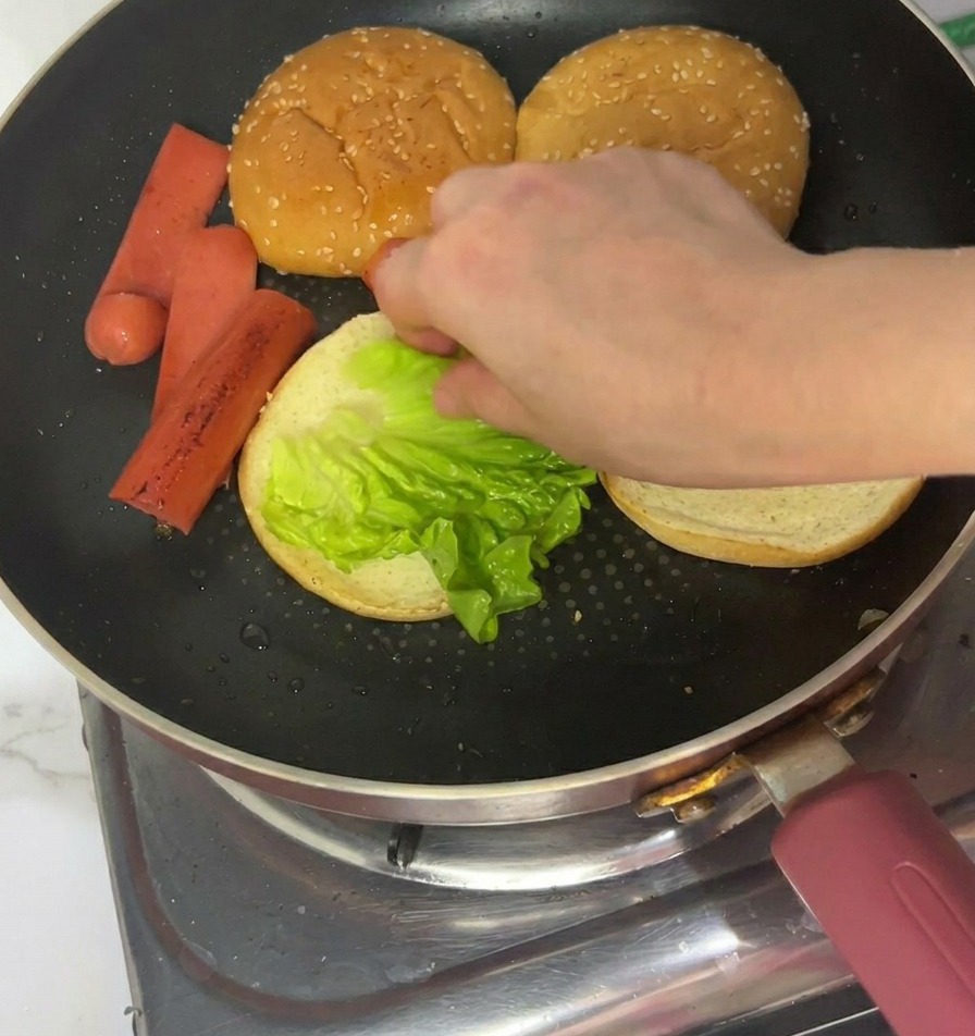 Una mano colocando una hoja de lechuga verde brillante sobre el lado tostado de un pan de hamburguesa en una sartén.