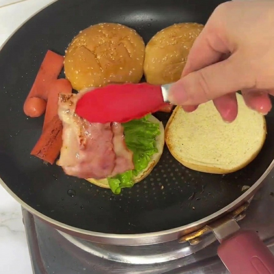 Pinzas rojas colocando una tira de tocino cocido y ondulado sobre un pan de hamburguesa con lechuga.