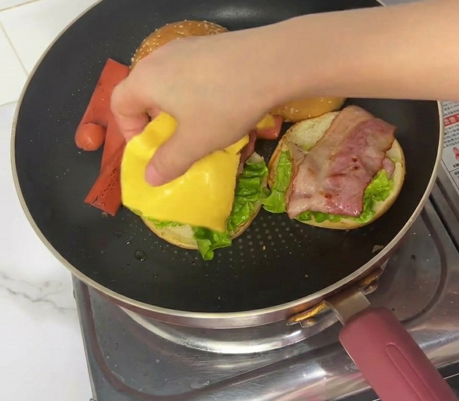 Una mano colocando una loncha cuadrada de queso procesado amarillo sobre el montaje de la hamburguesa en una sartén.