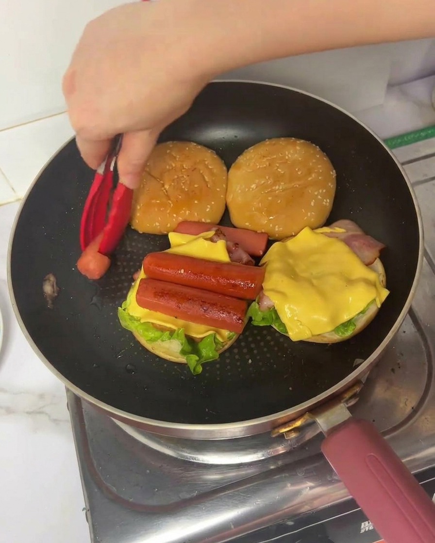 Una mano usando pinzas rojas para colocar un trozo doblado de huevo cocinado sobre una hamburguesa con lechuga, tocino, queso y salchichas.