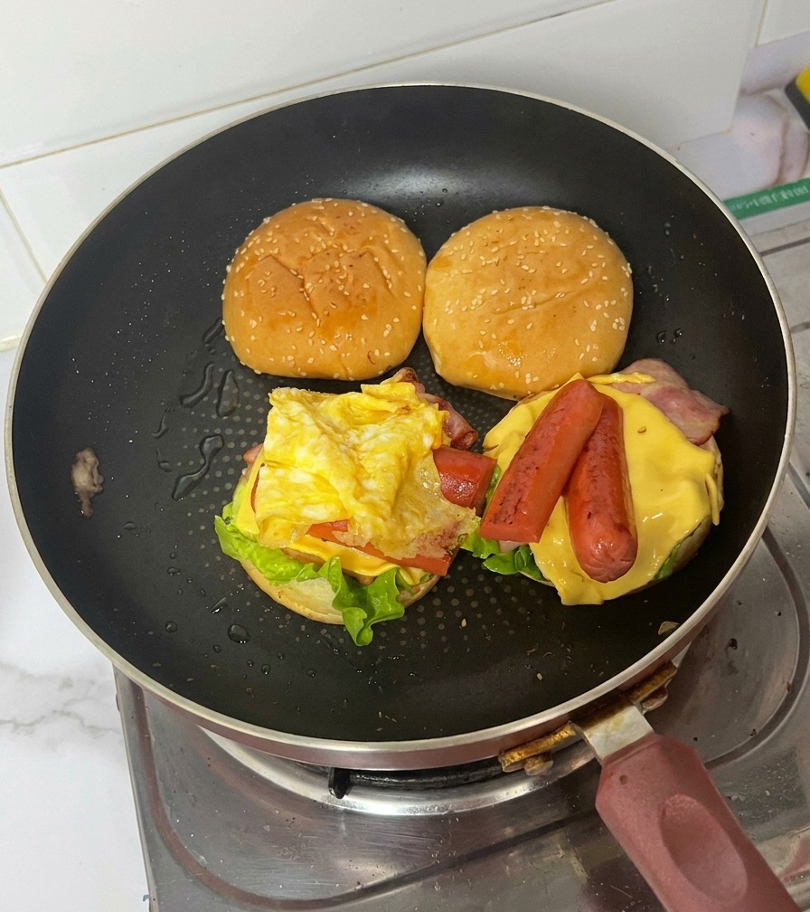 Montaje de hamburguesa en una sartén mostrando el huevo frito doblado sobre salchichas, queso, tocino y lechuga.