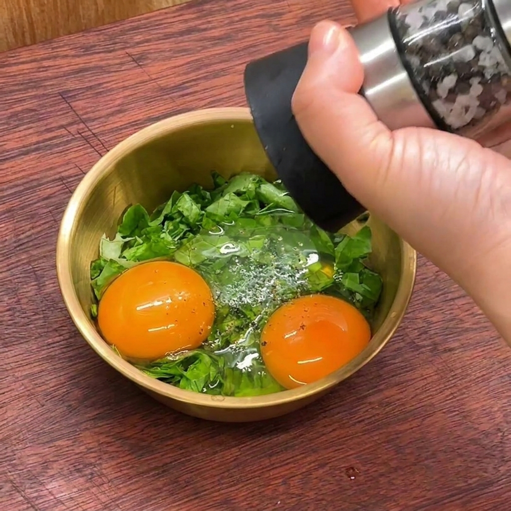 Una mano usando un molinillo de pimienta para sazonar yemas de huevo crudas y lechuga picada en un bol dorado.