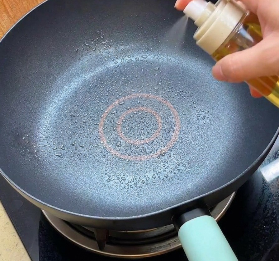 Una persona usando un pulverizador blanco para rociar aceite en una sartén antiadherente negra y limpia.