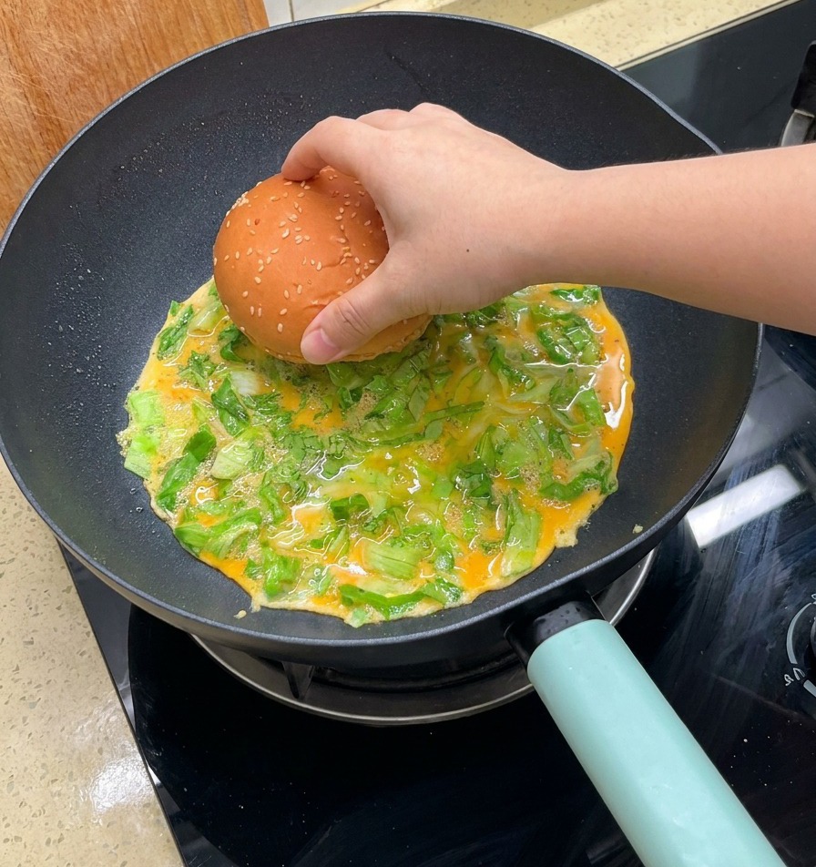 Una mano colocando un pan de sésamo sobre una tortilla de lechuga en cocción en la sartén.