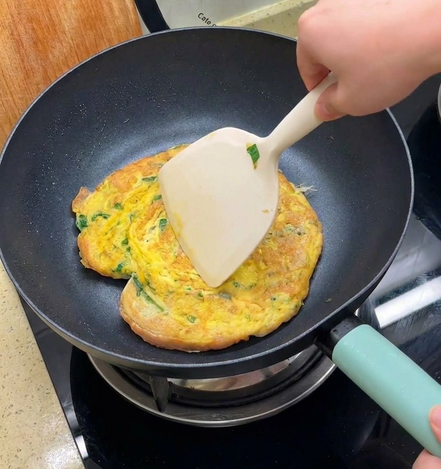 Una mano usando una espátula grande para dar la vuelta a una tortilla con pan en una sartén antiadherente negra.