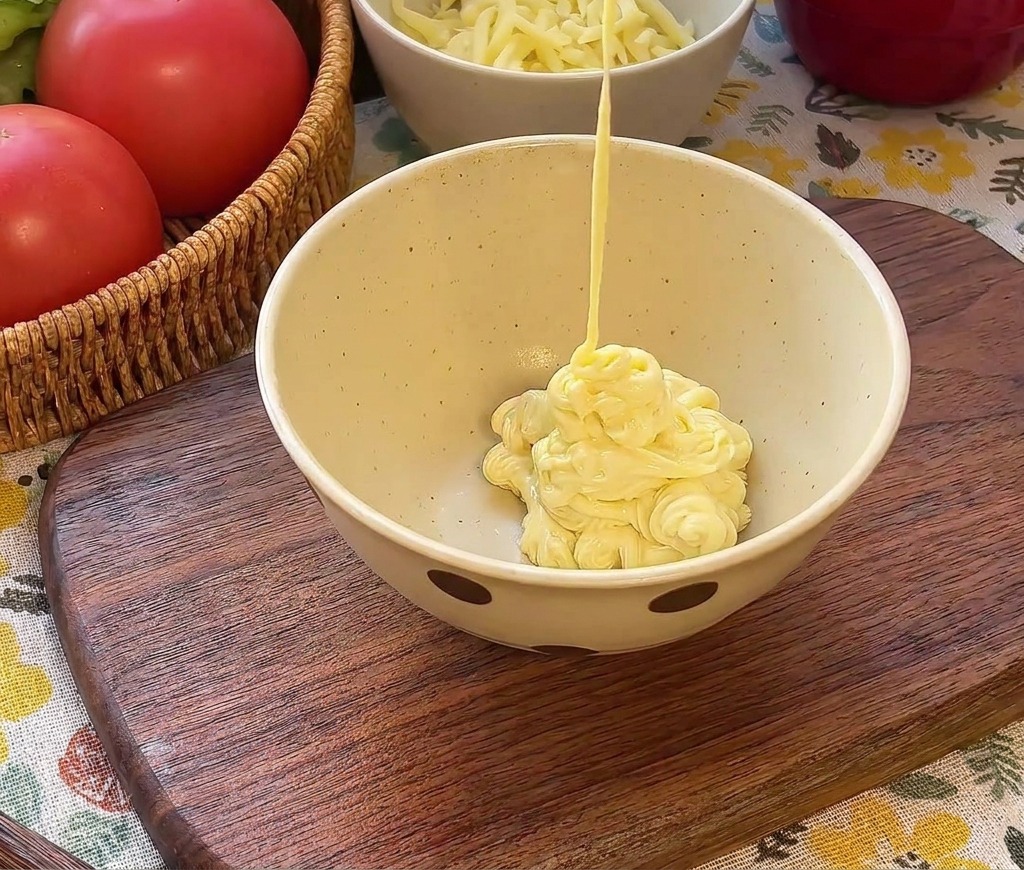 Mayonesa cremosa vertiéndose en un bol sobre kétchup y pepinillos picados.