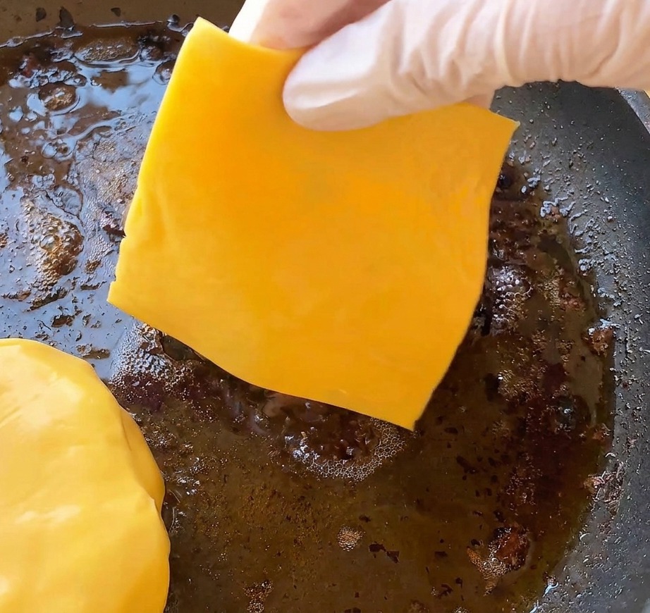 Una mano colocando una loncha de queso cheddar amarillo sobre la hamburguesa en la sartén.