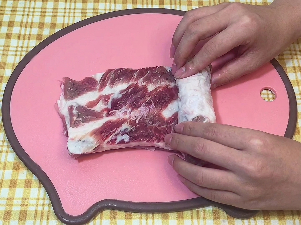 Manos enrollando firmemente un trozo de panceta cruda en forma de cilindro sobre una tabla de cortar rosa.