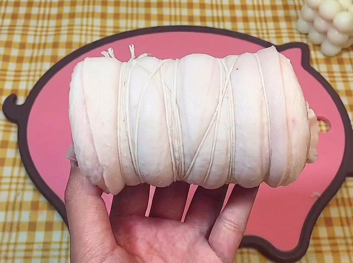 Una mano sosteniendo un rollo de panceta de cerdo cruda firmemente atado con hilo de cocina blanco.