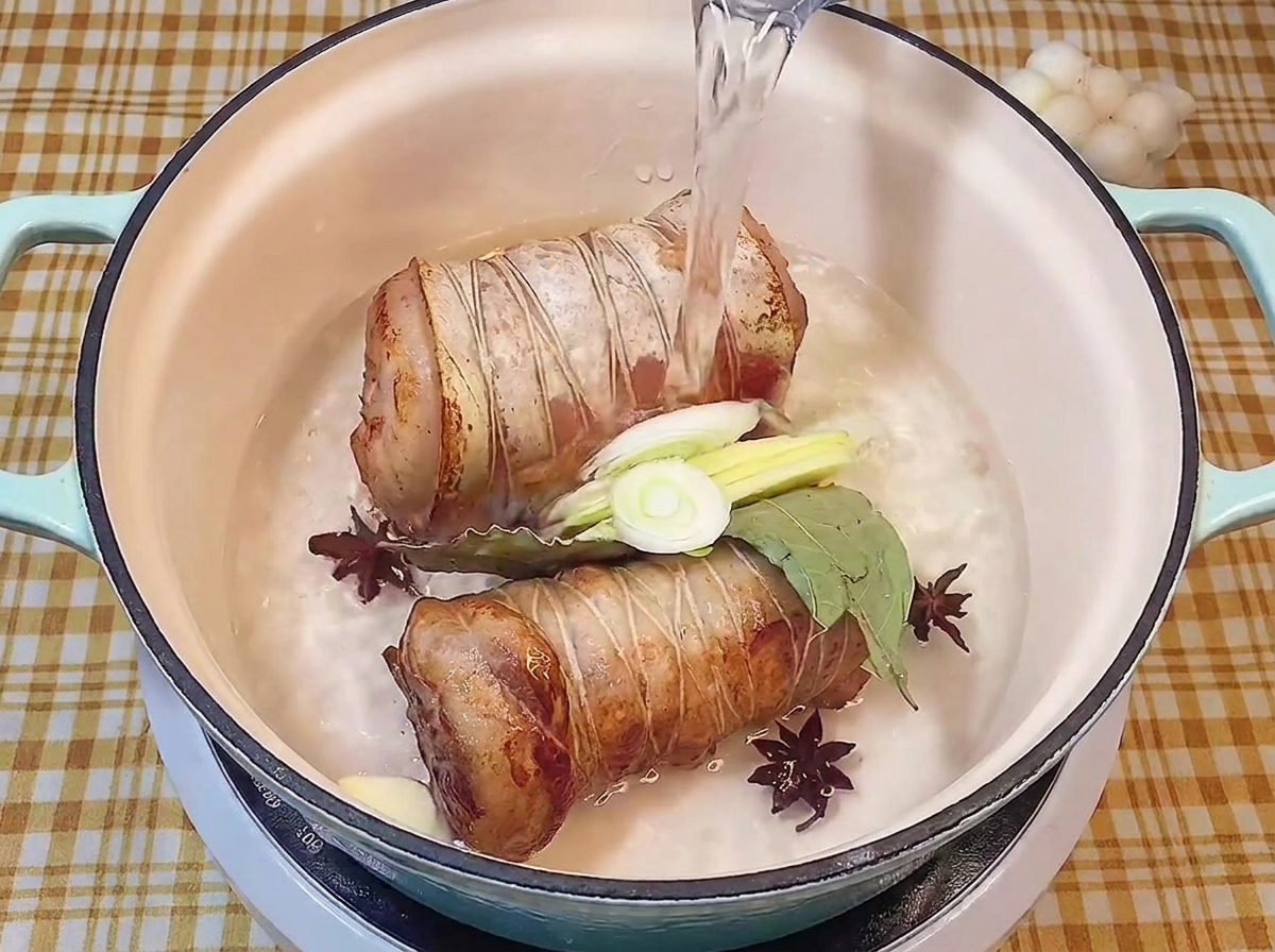 Vertiendo agua clara en una olla que contiene los rollos de panceta sellados junto con especias y aromáticos.